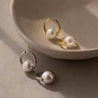 Two pairs of Bold Round Pearl Hoops—one gold, one silver—rest on a neutral ceramic dish, providing timeless style for lovers of statement earrings.