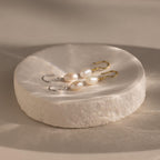 Two pairs of Dangling Pearl Diamond Earrings—one with gold hooks, one with silver—rest elegantly on a round white dish, perfect for fans of classic threader styles.