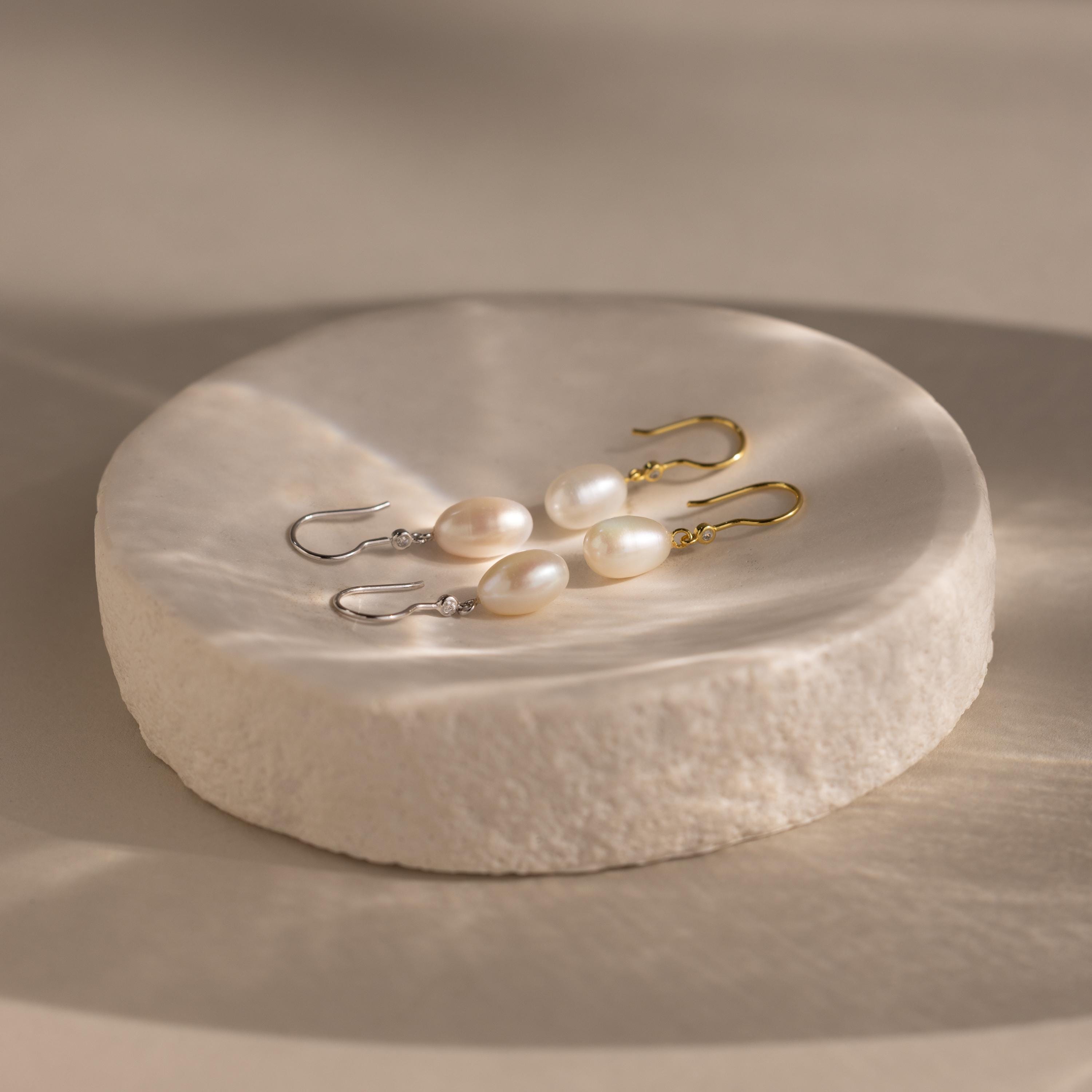 Two pairs of Dangling Pearl Diamond Earrings—one with gold hooks, one with silver—rest elegantly on a round white dish, perfect for fans of classic threader styles.
