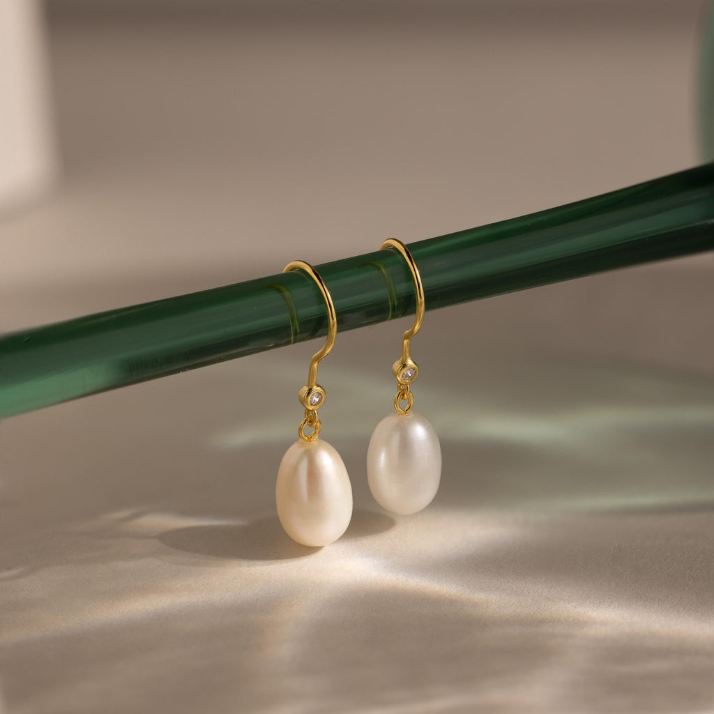 Dangling Pearl Diamond Earrings with white pearls and diamonds hang gracefully from a green glass rod against a neutral background.