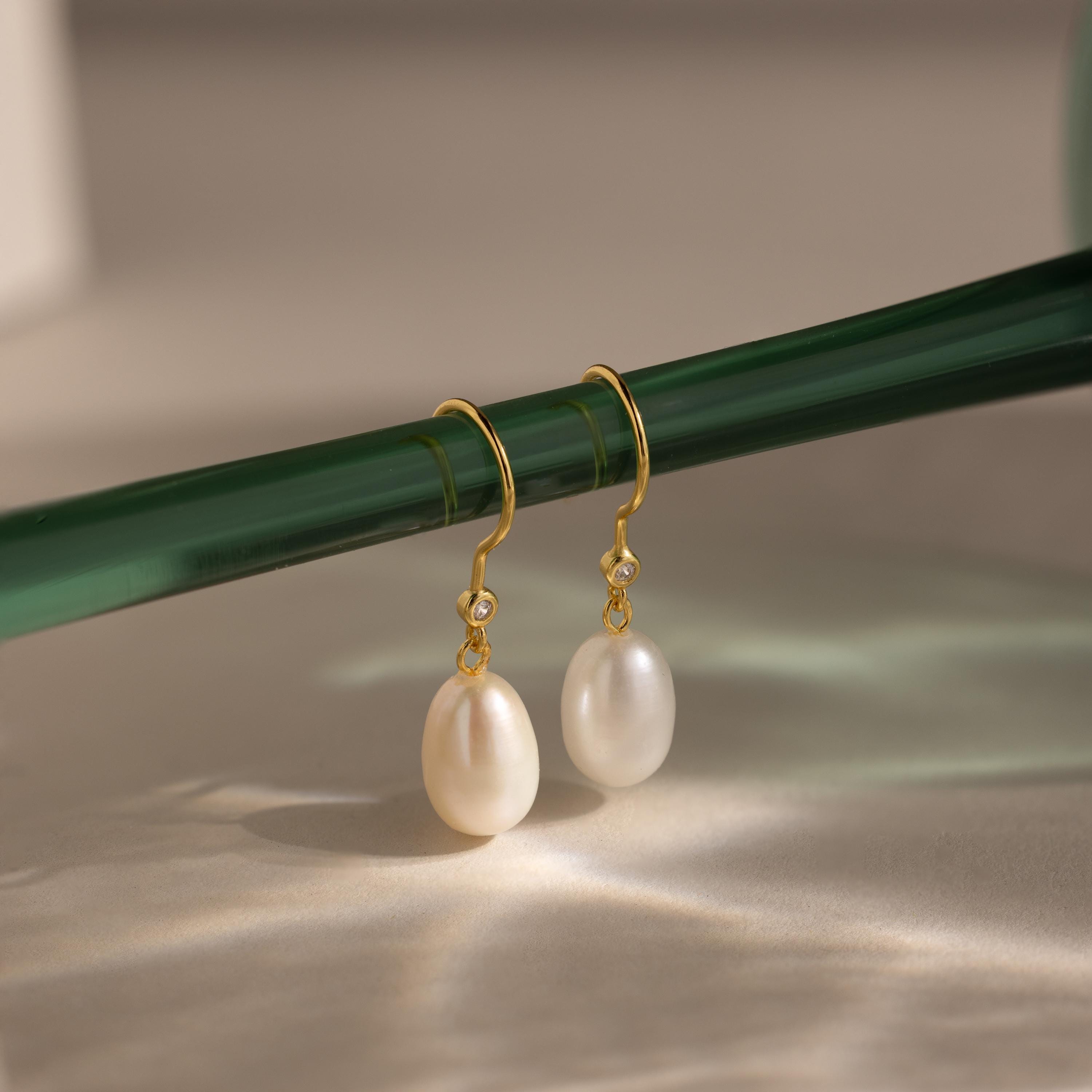 Dangling Pearl Diamond Earrings with white pearls and diamonds hang gracefully from a green glass rod against a neutral background.