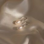 A pair of Dangling Pearl Diamond Earrings with silver hooks are displayed on soft, cream-colored satin fabric.