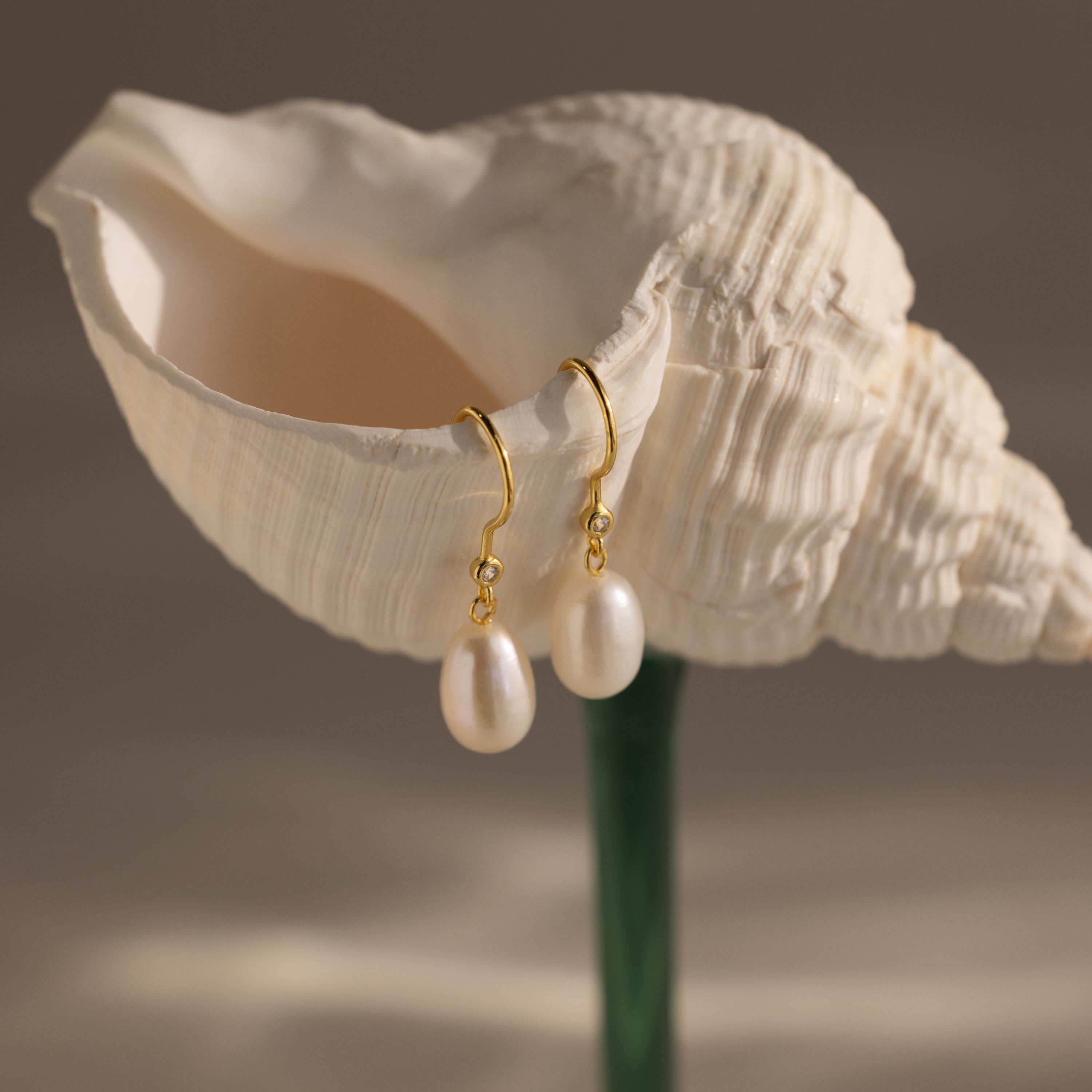 Dangling Pearl Diamond Earrings displayed hanging from a white seashell against a soft beige background.