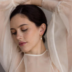 Woman wearing Dangling Pearl Diamond Earrings and a sheer cream blouse, eyes closed, with one arm raised behind her head.
