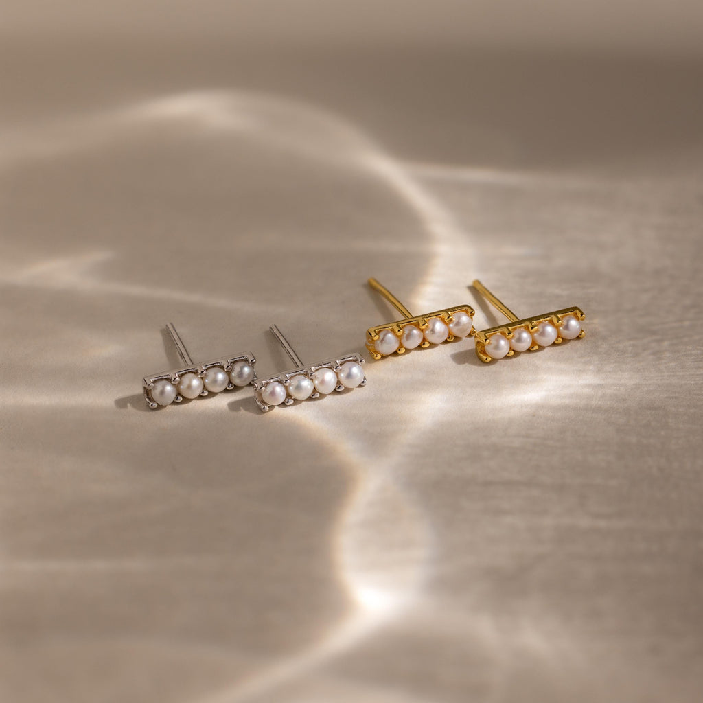 Four Dainty Pearl Bar Studs, featuring two silver and two gold designs, are displayed on a softly lit neutral surface.