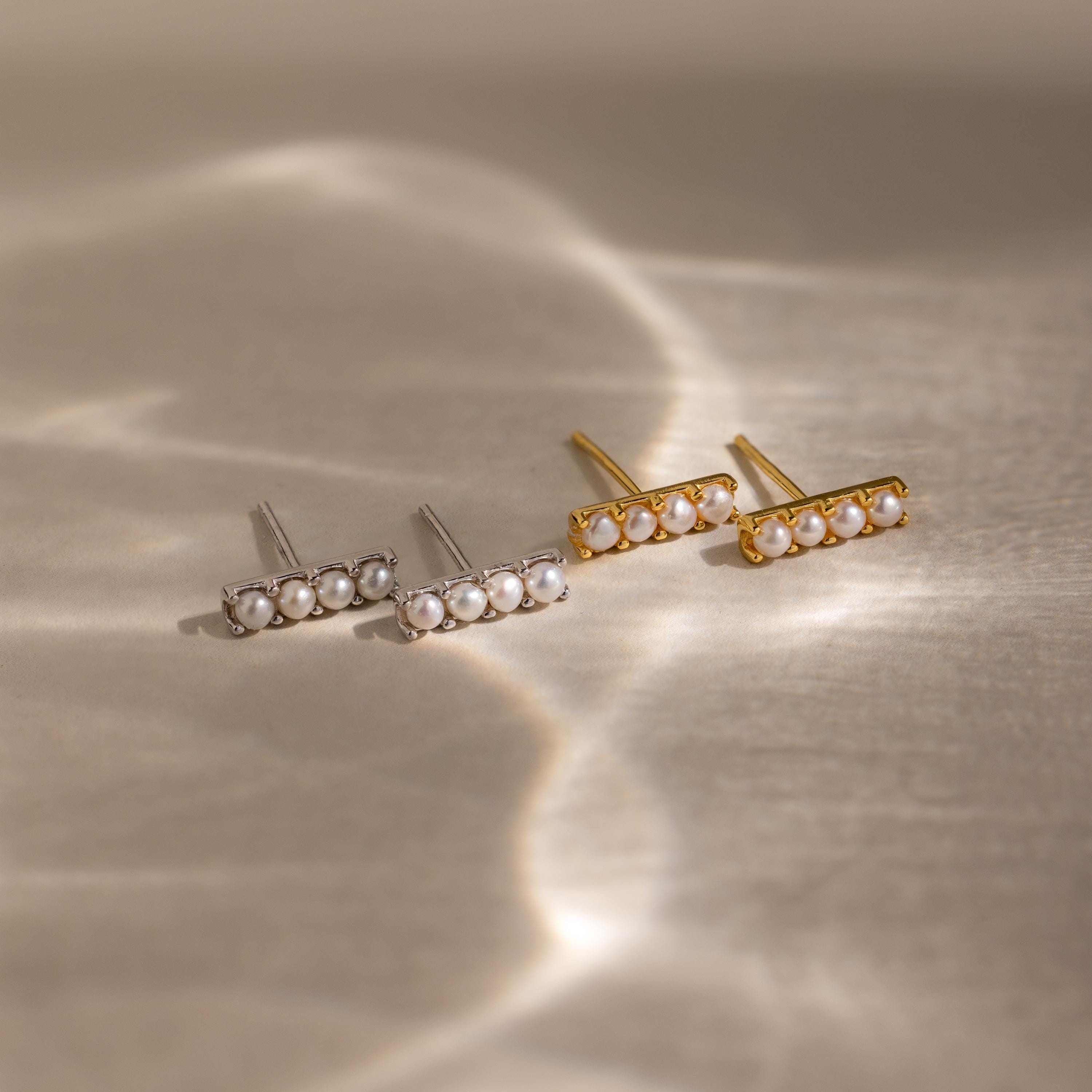 Four Dainty Pearl Bar Studs, featuring two silver and two gold designs, are displayed on a softly lit neutral surface.