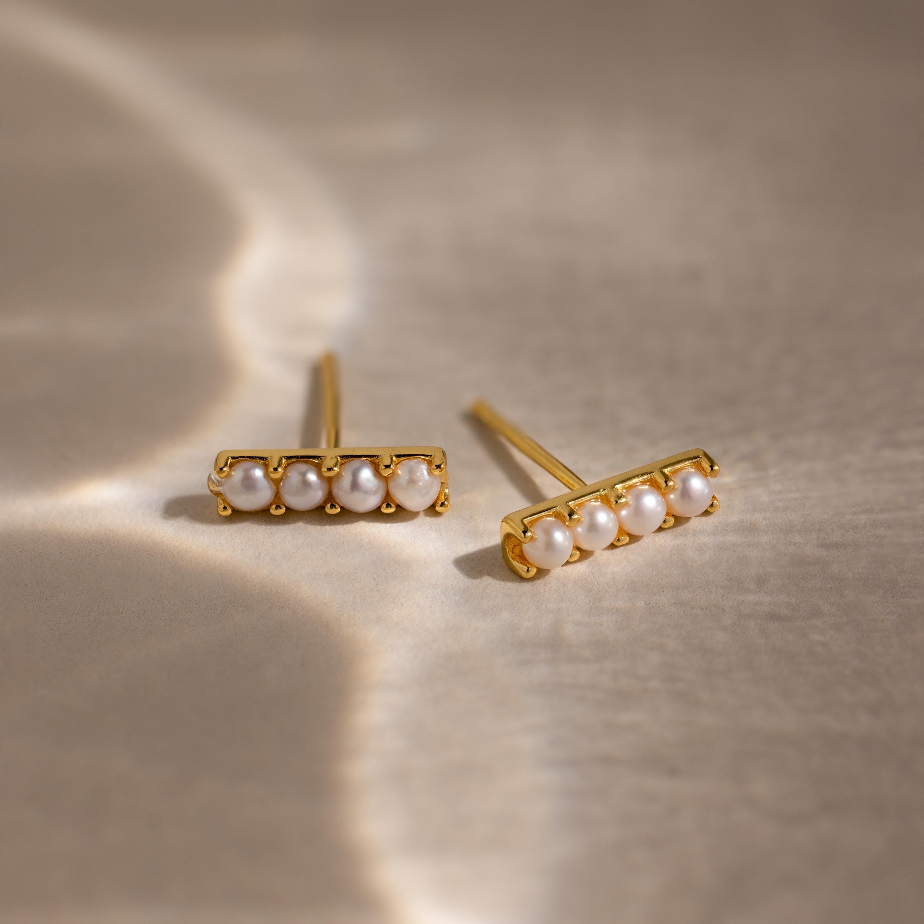 The Dainty Pearl Bar Studs feature four small pearls on gold bars, elegantly displayed on a softly lit beige surface.