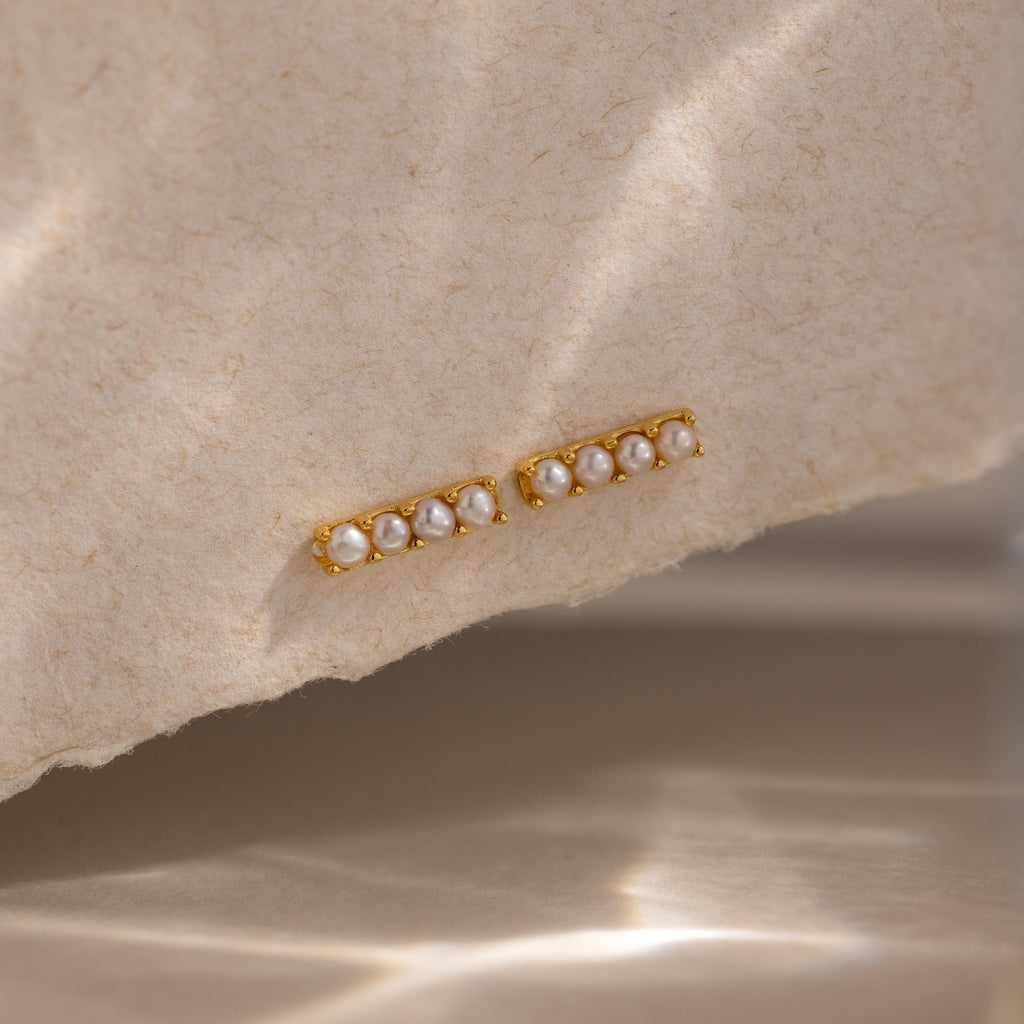 Dainty Pearl Bar Studs feature small pearls in a row on gold bars, adding subtle elegance to any look. Shown on a textured beige surface, they're perfect for those who love minimalist earrings.