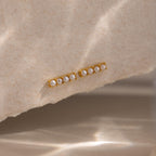 Dainty Pearl Bar Studs feature small pearls in a row on gold bars, adding subtle elegance to any look. Shown on a textured beige surface, they're perfect for those who love minimalist earrings.