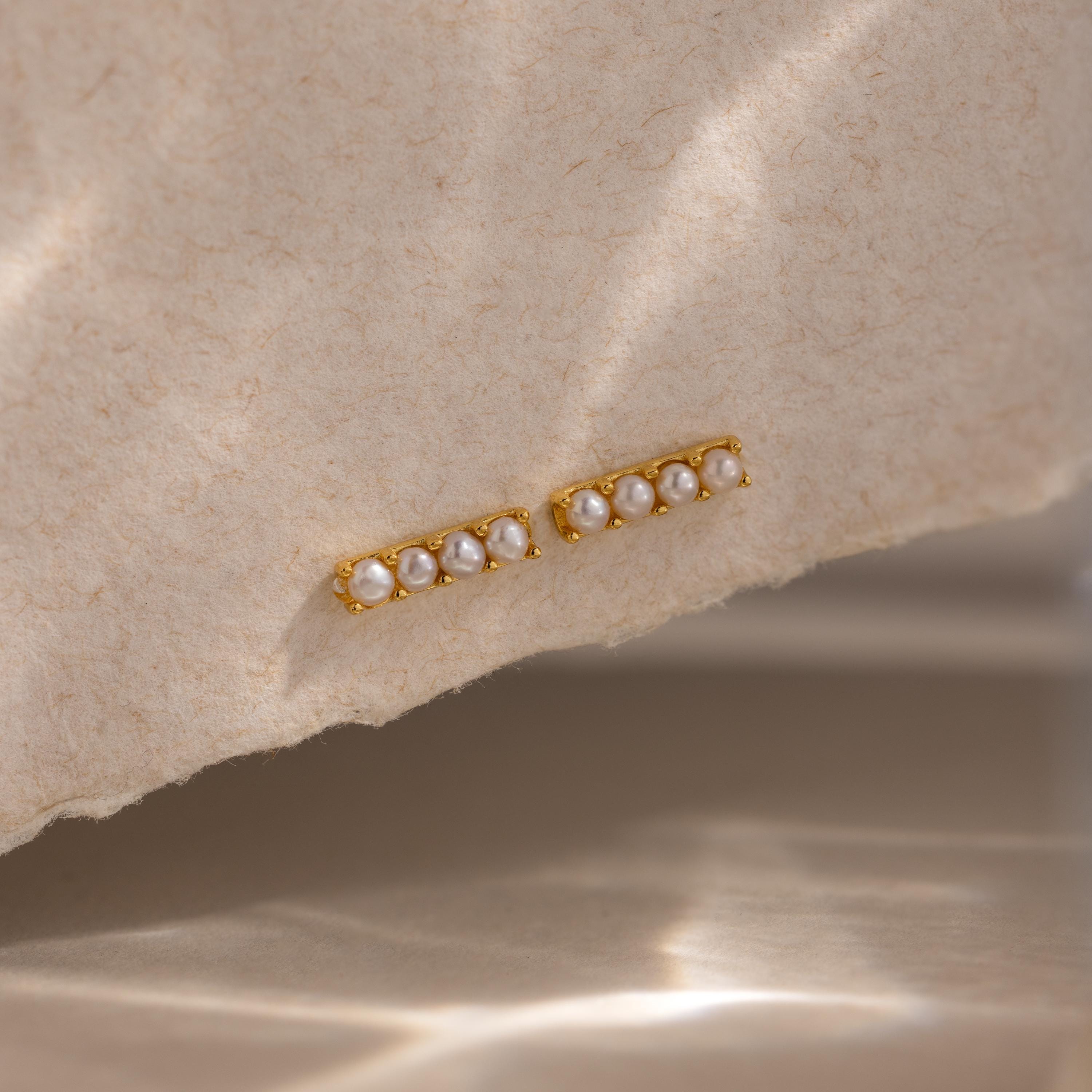 Dainty Pearl Bar Studs feature small pearls in a row on gold bars, adding subtle elegance to any look. Shown on a textured beige surface, they're perfect for those who love minimalist earrings.