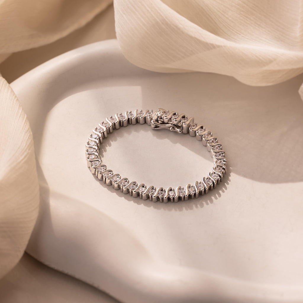 The S-Link Diamond Tennis Bracelet, featuring shimmering stones, is displayed on a white ceramic surface and accented with soft fabric for an elegant touch.