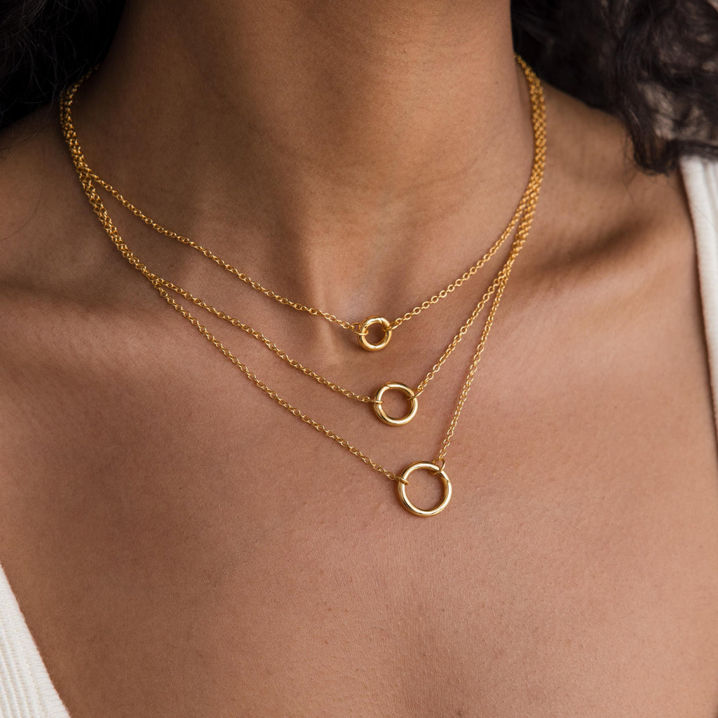 A woman wears the Round Carabiner Charm Set in 18K Gold, featuring three gold chain necklaces with small, medium, and large circle pendants styled as a charm necklace.