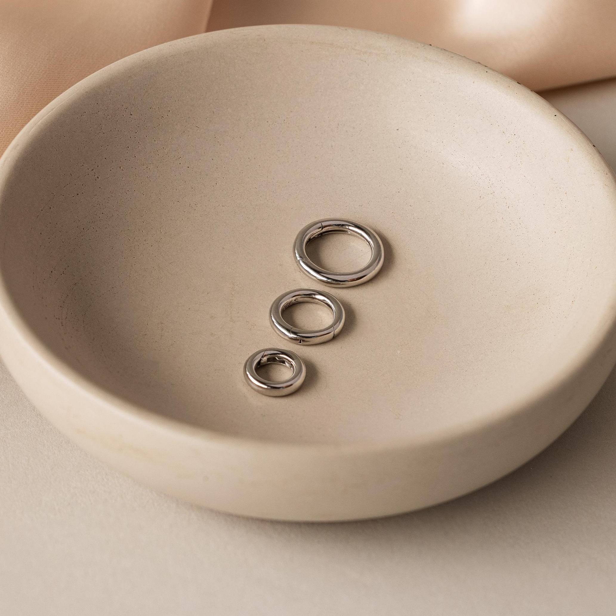 Three sterling silver rings in varying sizes rest in a round beige ceramic dish—ideal accents for your charm bracelet or to pair with the Round Carabiner Charm Set in Sterling Silver.