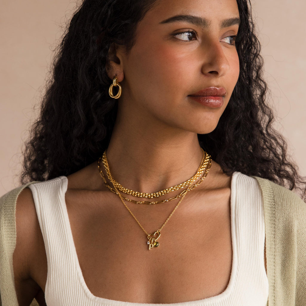 Woman with long curly hair wears the Heart Carabiner Set in 18K Gold—necklaces, charm bracelet connector, and hoop earrings—paired with a white top and a light green cardigan.