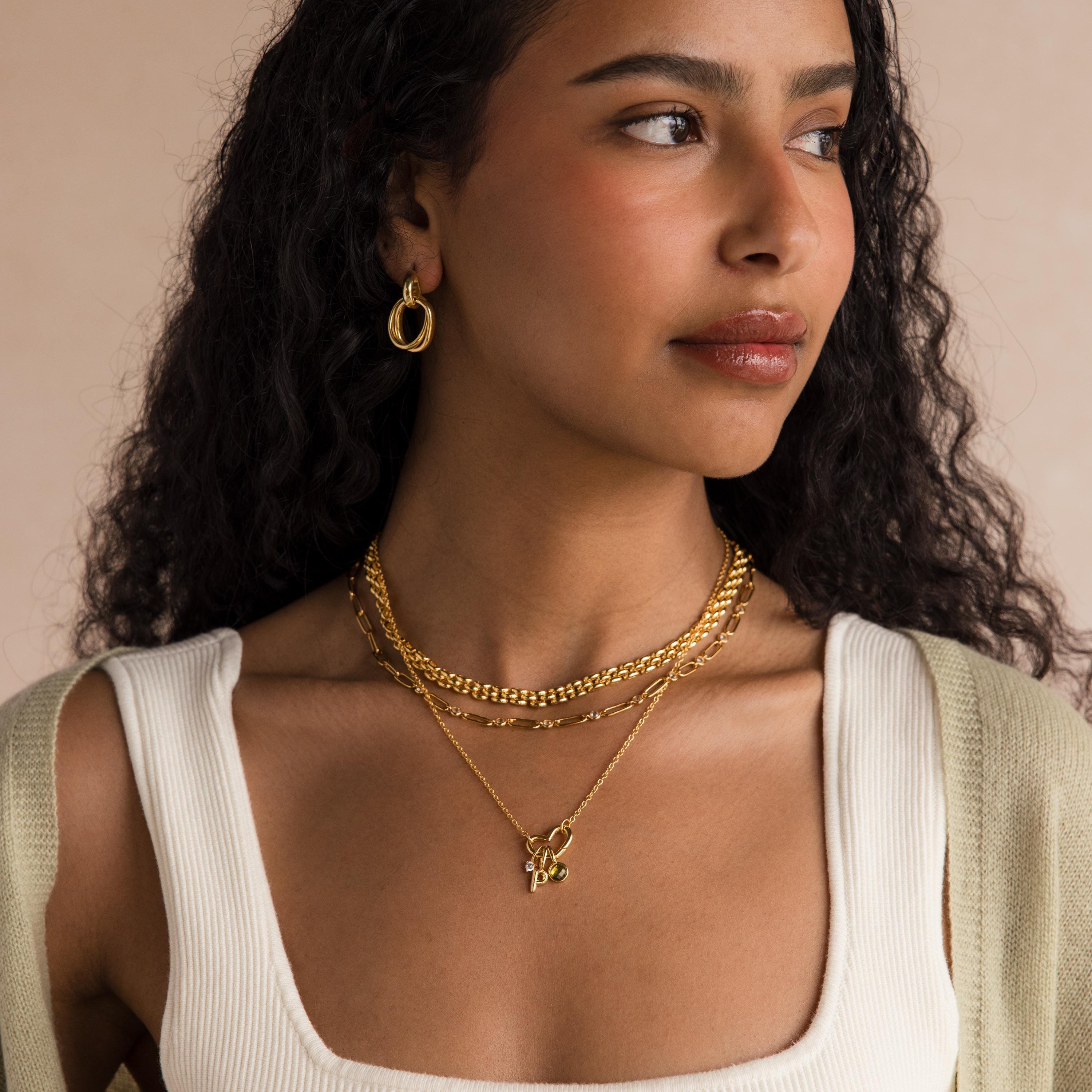 Woman with long curly hair wears the Heart Carabiner Set in 18K Gold—necklaces, charm bracelet connector, and hoop earrings—paired with a white top and a light green cardigan.