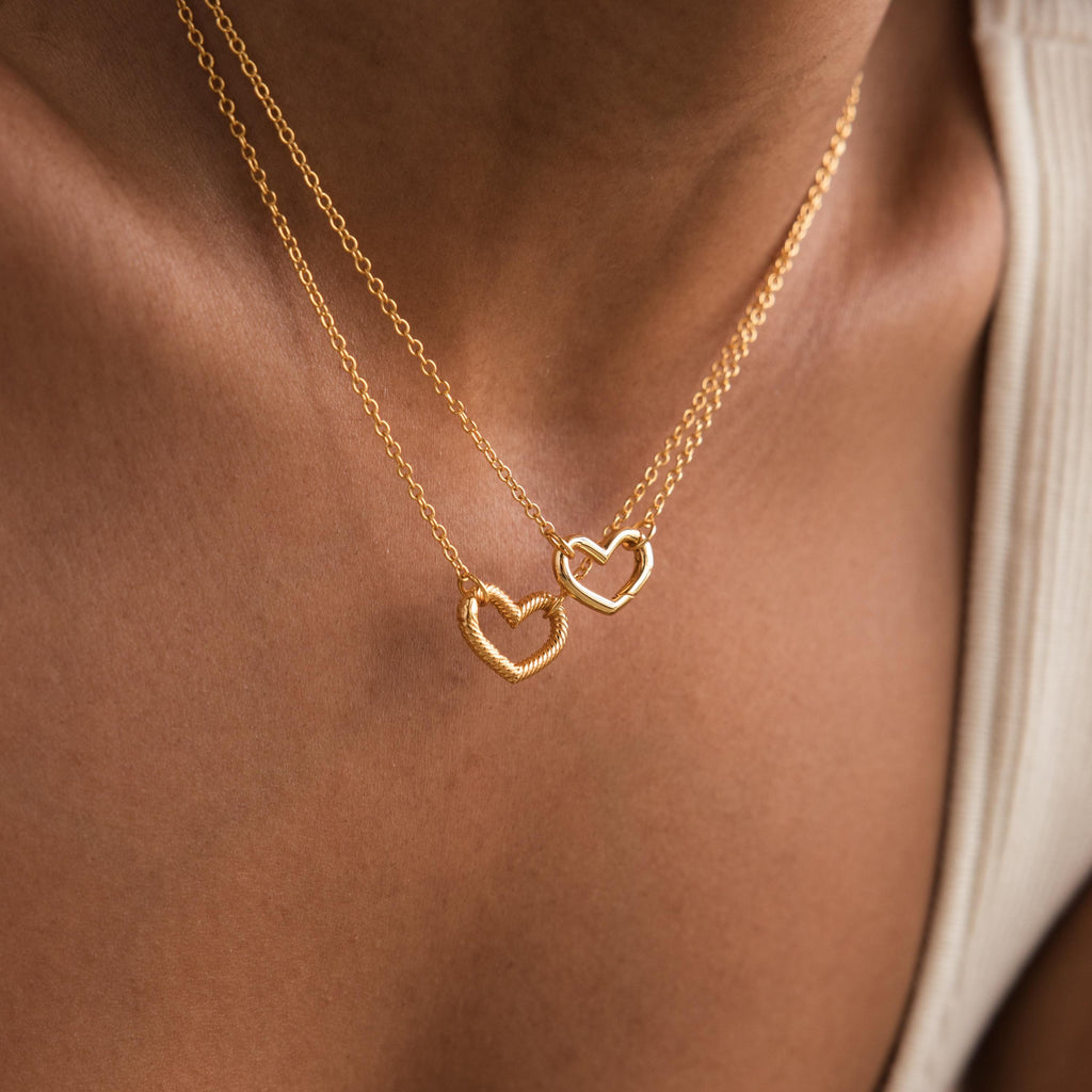 Two Heart Carabiner Set in 18K Gold necklaces rest on a person's neck, each with a delicate heart-shaped carabiner. The close-up captures the jewelry's charm and elegance against the skin, highlighting this stunning set.