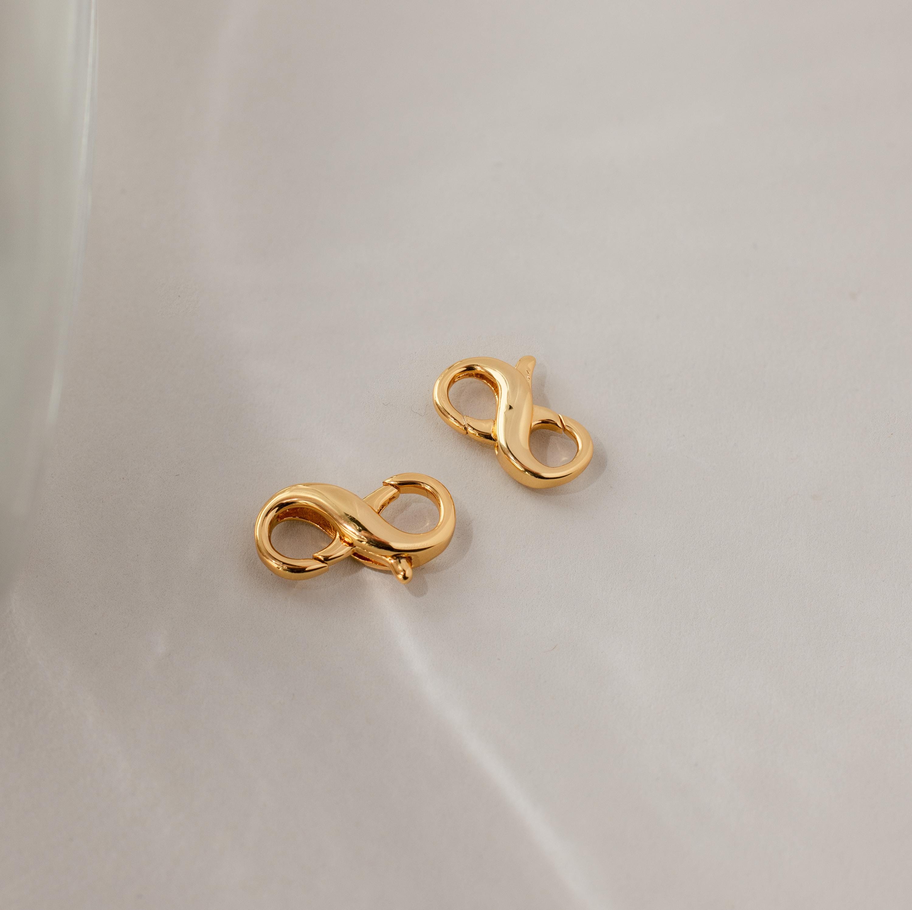 The Multipurpose Lobster Clasp Extender Set in 18K Gold, shaped in an infinity design, rests on a light, textured surface—ideal for use as a bracelet or necklace extender.