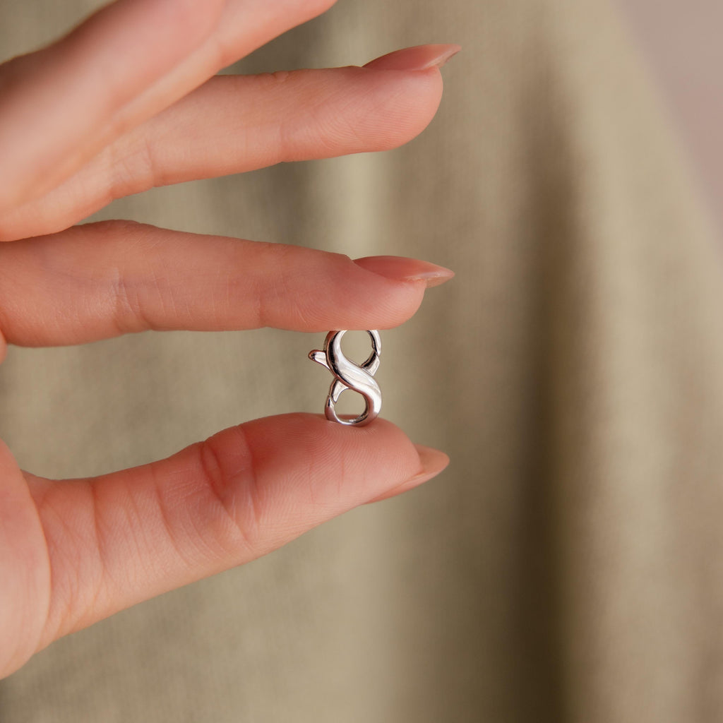 A manicured hand holds a Multipurpose Lobster Clasp Extender Set in White Gold, featuring a small infinity-shaped charm, against a beige background.