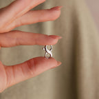A manicured hand holds a Multipurpose Lobster Clasp Extender Set in White Gold, featuring a small infinity-shaped charm, against a beige background.