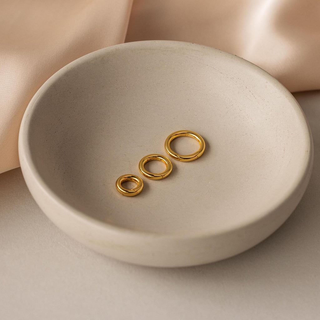 The Round Carabiner Charm Set in 18K Gold features three gold rings in varying sizes, ideal for charm necklaces or bracelets, pictured in a round ceramic dish on a soft beige background.