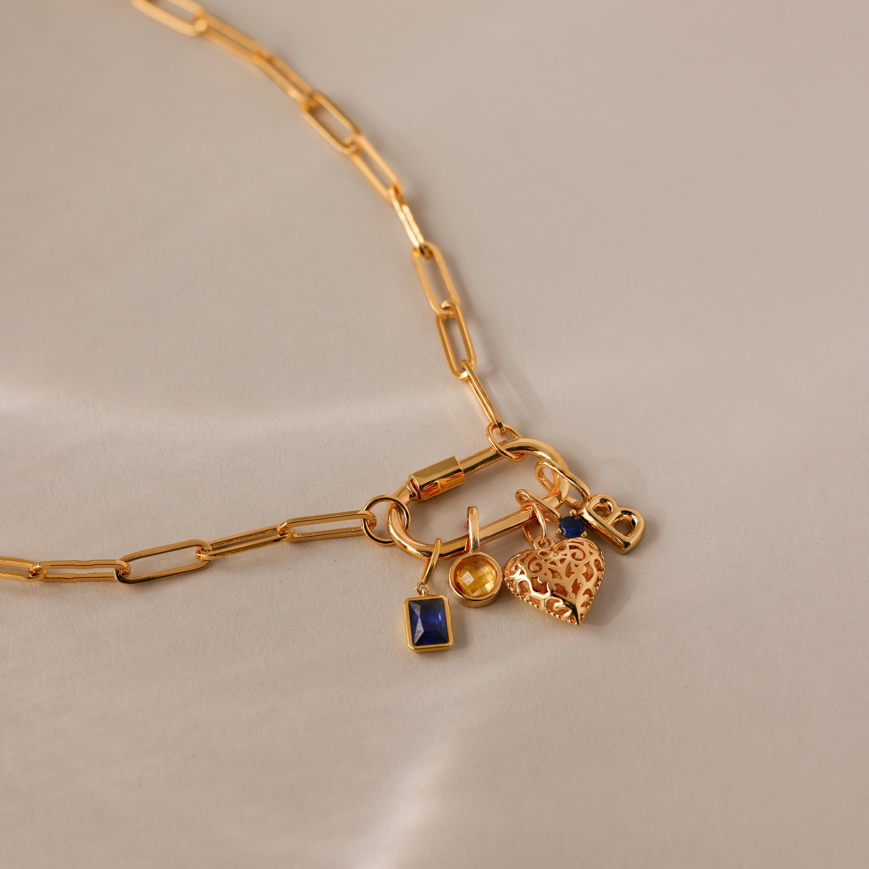 The Bar & Carabiner Charm Set includes a gold chain necklace with heart, blue square, yellow circle, and small lock charms plus charm connectors, displayed on a beige background.