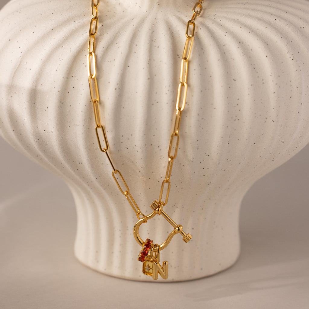 Bar & Carabiner Charm Set in 18K Gold, with a red gemstone charm and N pendant, secured by a carabiner clasp, displayed draped over a white ceramic vase.