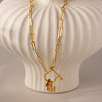 Bar & Carabiner Charm Set in 18K Gold, with a red gemstone charm and N pendant, secured by a carabiner clasp, displayed draped over a white ceramic vase.