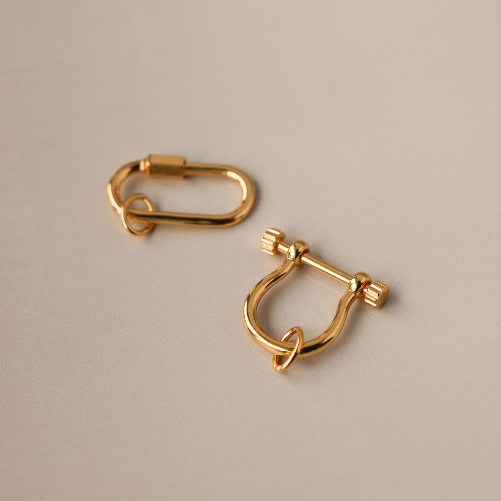 Two gold charm connectors—one oval and one U-shaped with a bar—rest on a smooth beige surface, perfect as necklace accessories or as part of the Bar & Carabiner Charm Set.