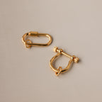 Two gold charm connectors—one oval and one U-shaped with a bar—rest on a smooth beige surface, perfect as necklace accessories or as part of the Bar & Carabiner Charm Set.