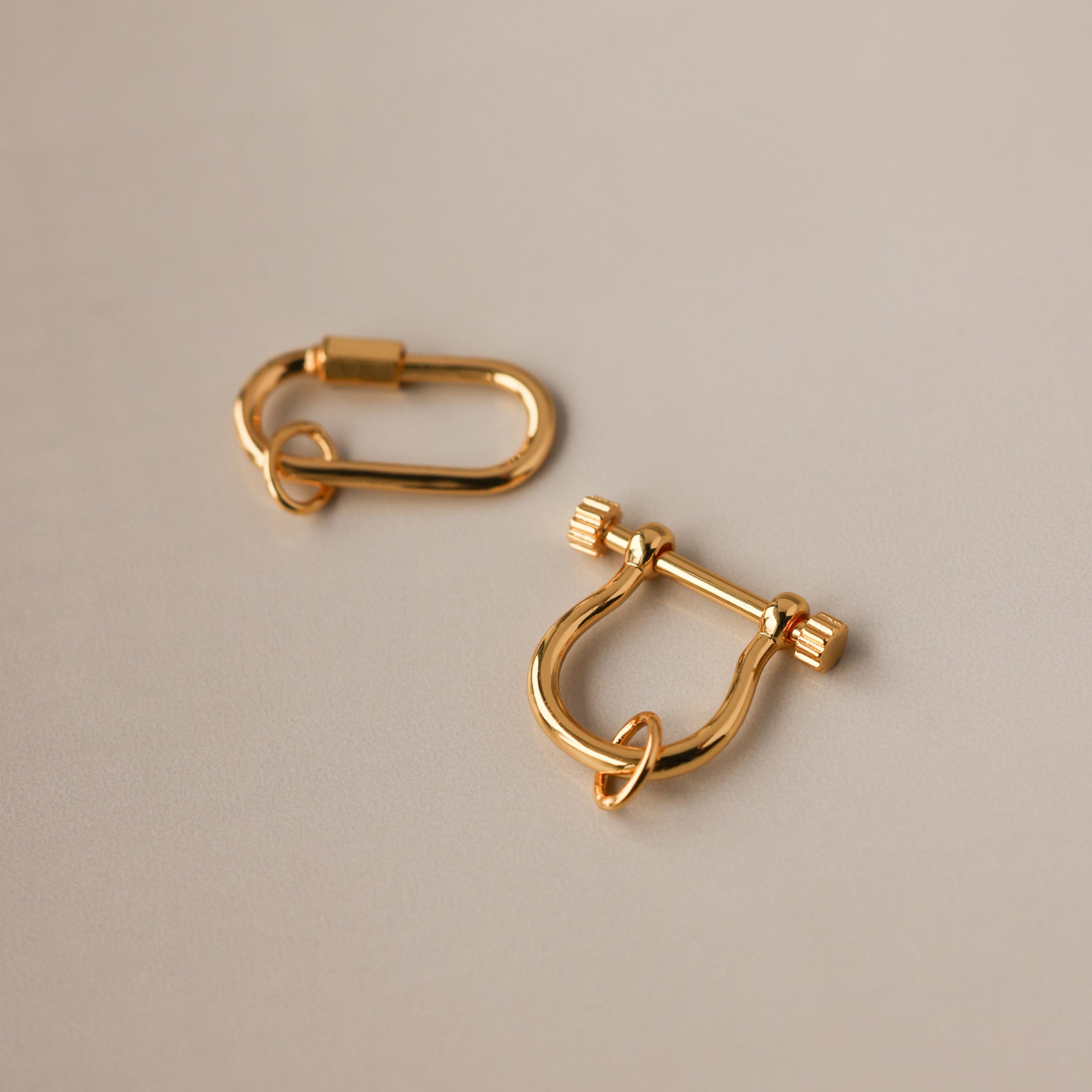 Two gold charm connectors—one oval and one U-shaped with a bar—rest on a smooth beige surface, perfect as necklace accessories or as part of the Bar & Carabiner Charm Set.