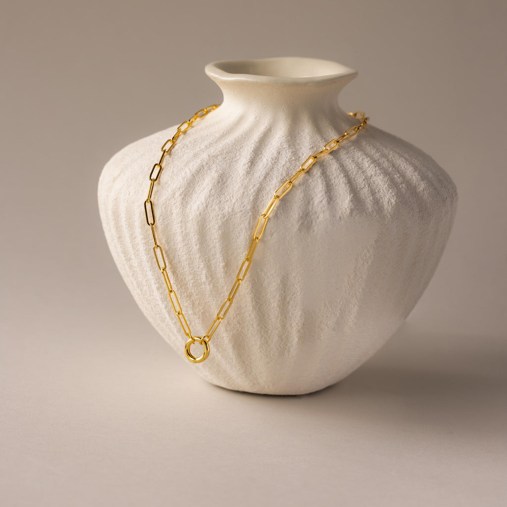 Paperclip Clasp Necklace draped over a textured white ceramic vase on a neutral background, featuring collectible charms for a unique touch.