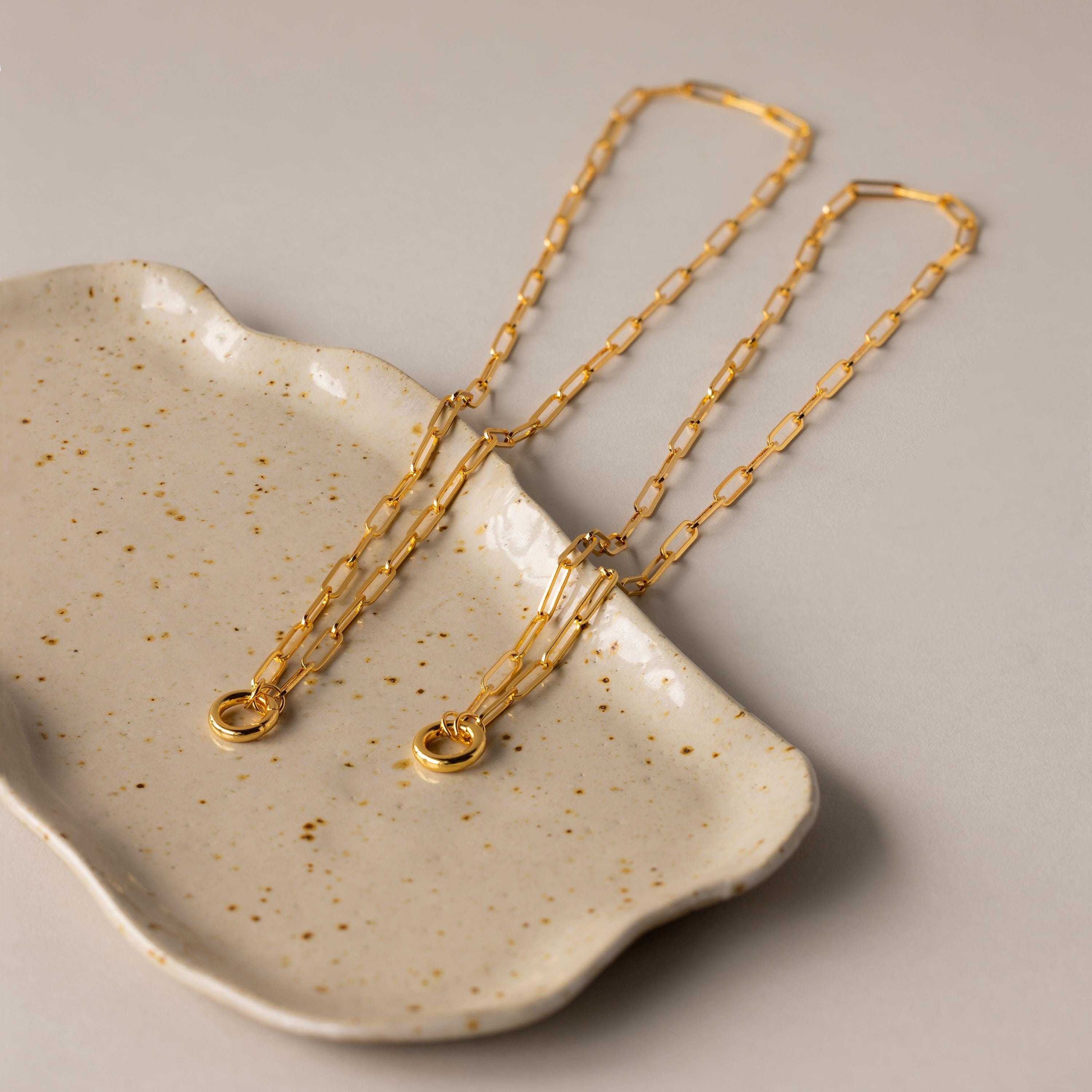 The Paperclip Clasp Necklace, a striking gold chain, is elegantly displayed on a speckled ceramic dish set against a beige background.