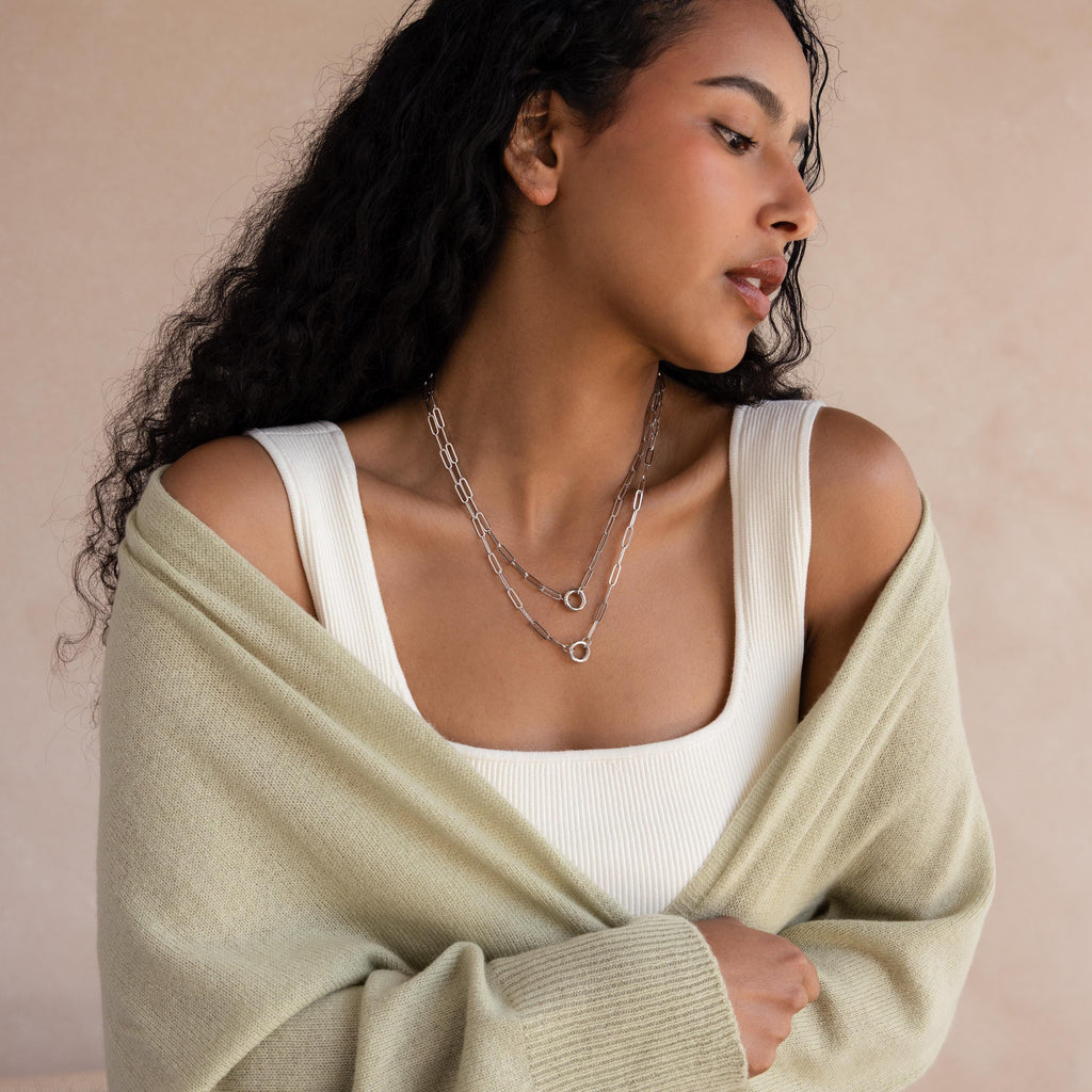 Woman with long curly hair wearing a Paperclip Clasp Necklace in White Gold, layered silver necklaces, a white top, and a light green cardigan.