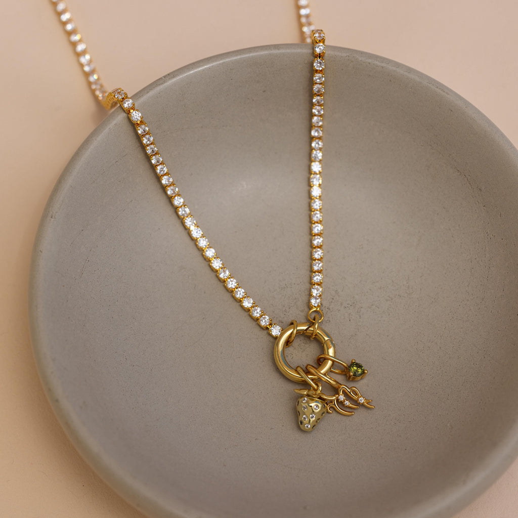 The Diamond Tennis Clasp Necklace in 18K Gold shines with playful charm details, including a strawberry, and features a secure rolo clasp—perfect for adding elegance to your wedding jewelry collection. Displayed on a round gray dish.