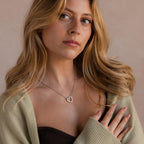 A woman with long blonde hair and a light sweater looks thoughtful, her outfit accented by the Rolo Heart Clasp Necklace in White Gold, whose heart-shaped clasp adds a subtle touch of elegance.
