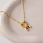 The Rolo Heart Clasp Necklace in 18K Gold features a gold heart pendant, purple gemstone charm, and letter B accent, displayed on a beige ceramic dish with a stylish rolo chain and heart-shaped clasp.