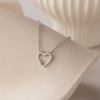 The Rolo Heart Clasp Necklace features a silver heart-shaped pendant on a delicate rolo chain, shown on a smooth, light-colored curved surface with a soft background.