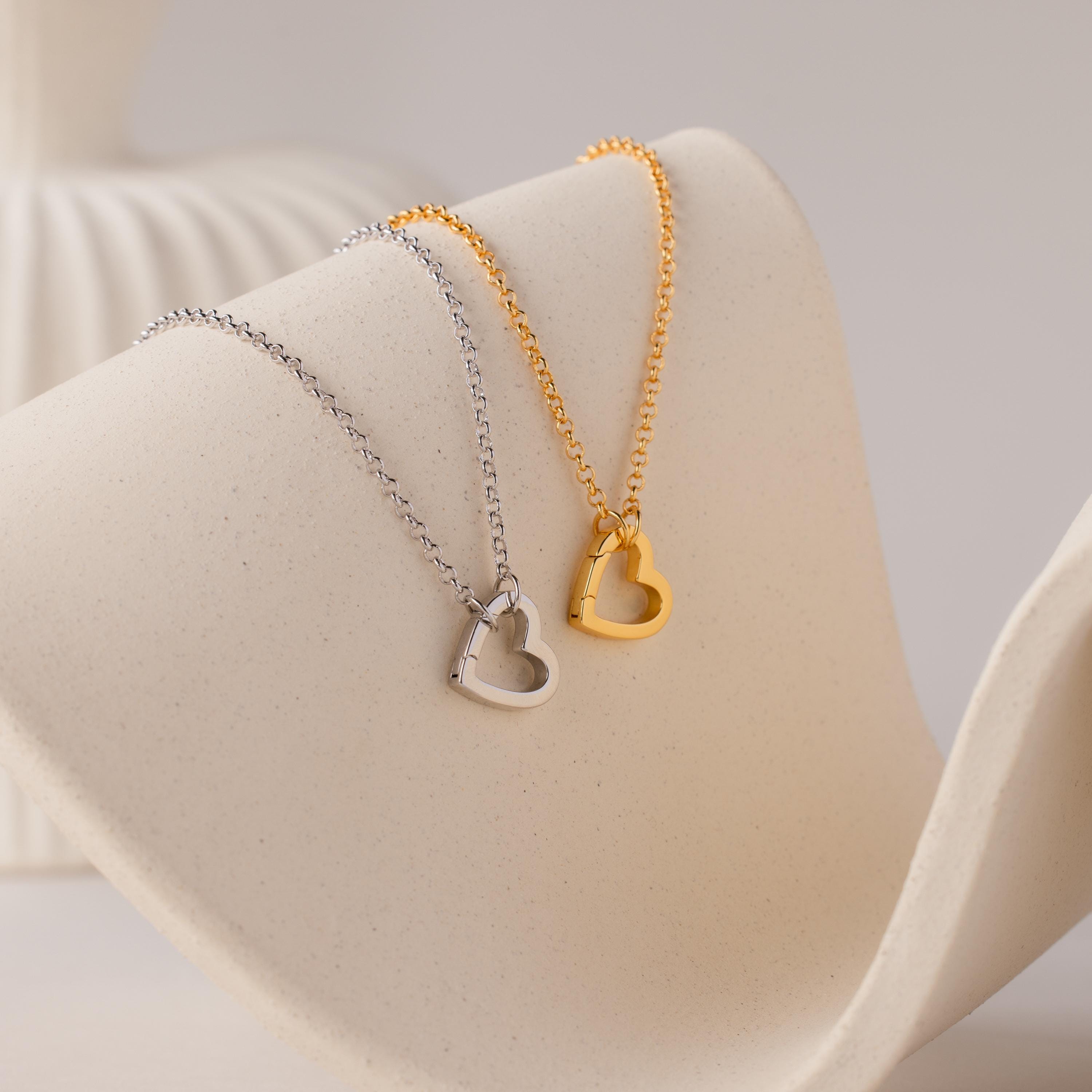 The Rolo Heart Clasp Necklace features a delicate rolo chain and a heart-shaped pendant, available in silver or gold, elegantly displayed on a curved cream-colored surface.