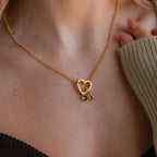 A woman wears the Rolo Heart Clasp Necklace featuring a gold heart, purple gem, and letter B charm on a delicate rolo chain, styled over a cozy sweater and top.