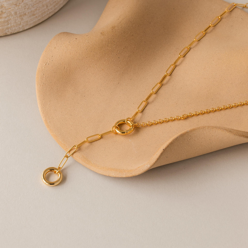 The Rolo Lariat Paperclip Clasp Necklace in 18K Gold, featuring circular links and a lariat clasp, is displayed on a curved beige ceramic surface.