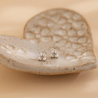 The Pearl Ivy Flower Studs, delicate silver wedding earrings, are displayed on a textured ceramic heart-shaped dish against a soft beige background.