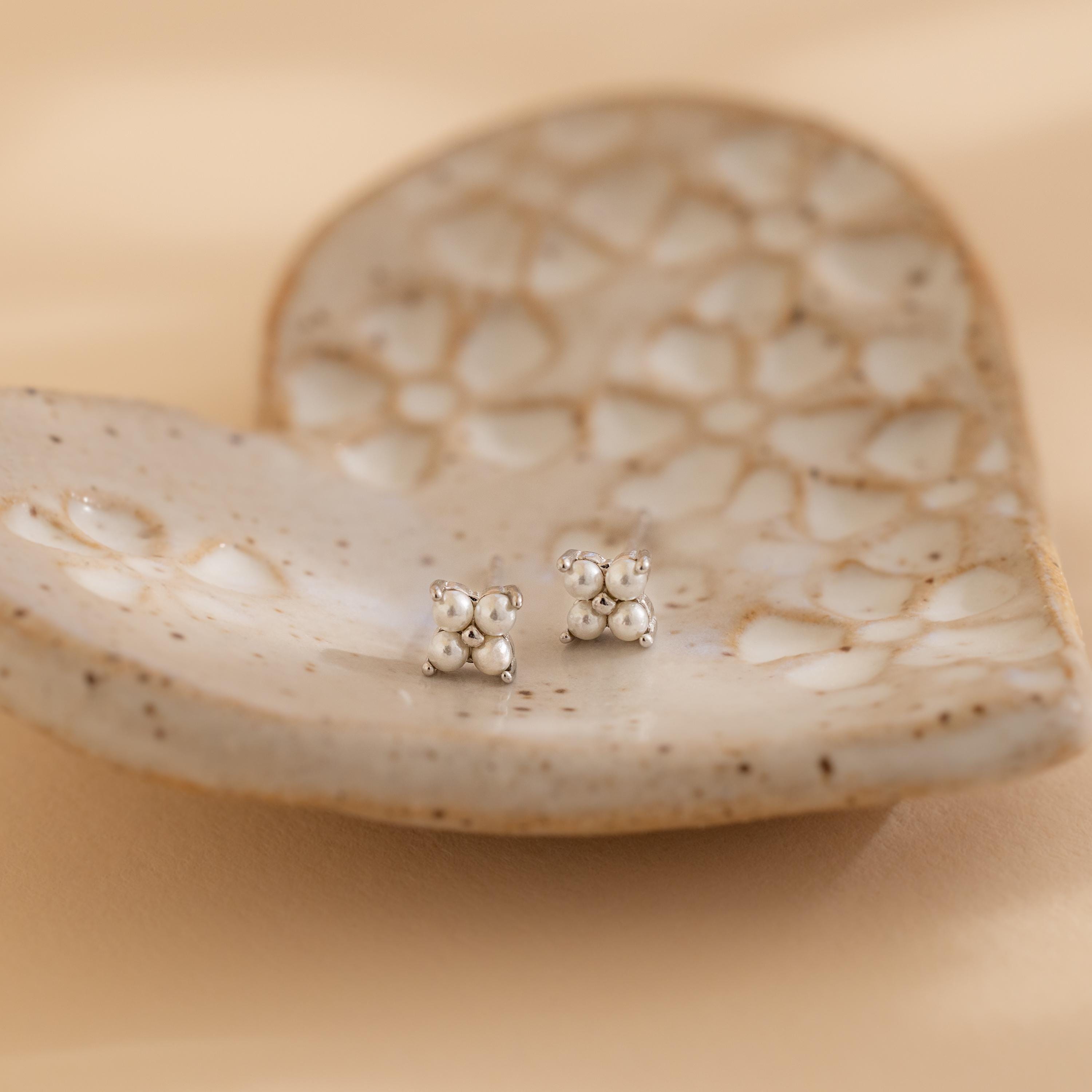 The Pearl Ivy Flower Studs, delicate silver wedding earrings, are displayed on a textured ceramic heart-shaped dish against a soft beige background.