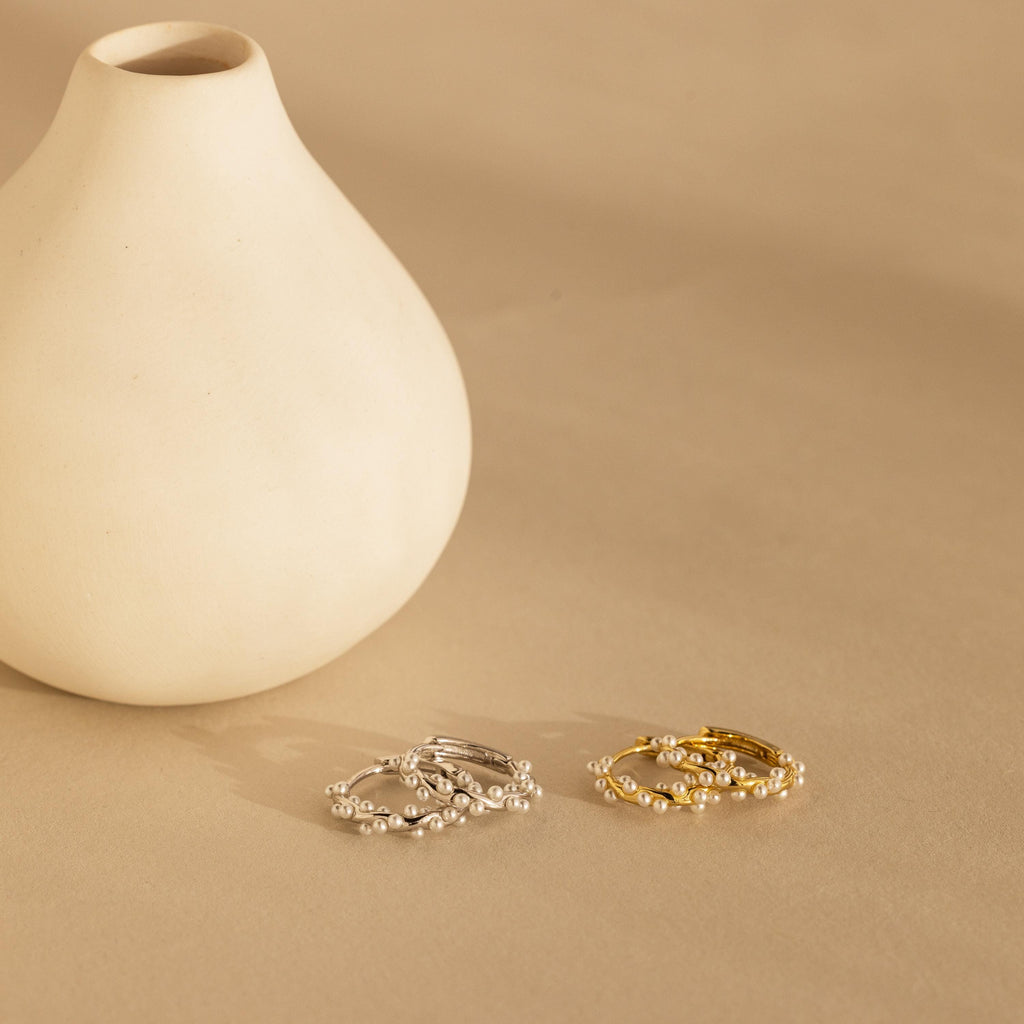 Minimalist ceramic vase with two sets of pearl-accented rings, inspired by bridal earrings and the Pearl Cluster Huggies in 18K Gold, one silver and one gold, elegantly displayed on a beige surface.