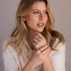 A woman with long blonde hair wears a cream sweater and showcases the Dainty Marquise Topaz Ring, styled with gold and red rings, while glancing to the side against a neutral backdrop.