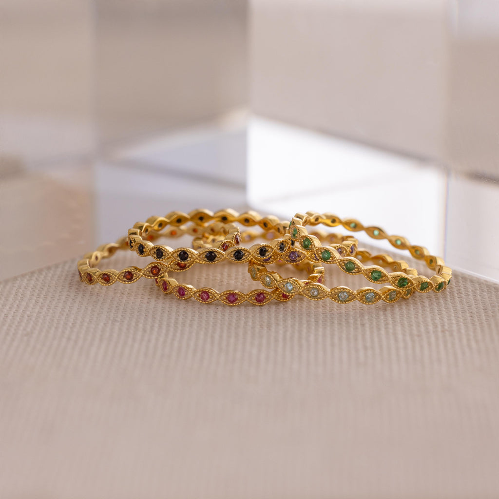 A dainty Marquise Birthstone Ring is paired with gold bangles set with colorful gemstones, all arranged on a textured beige surface near clear acrylic blocks for a touch of personalized elegance.