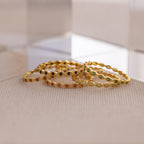 A dainty Marquise Birthstone Ring is paired with gold bangles set with colorful gemstones, all arranged on a textured beige surface near clear acrylic blocks for a touch of personalized elegance.