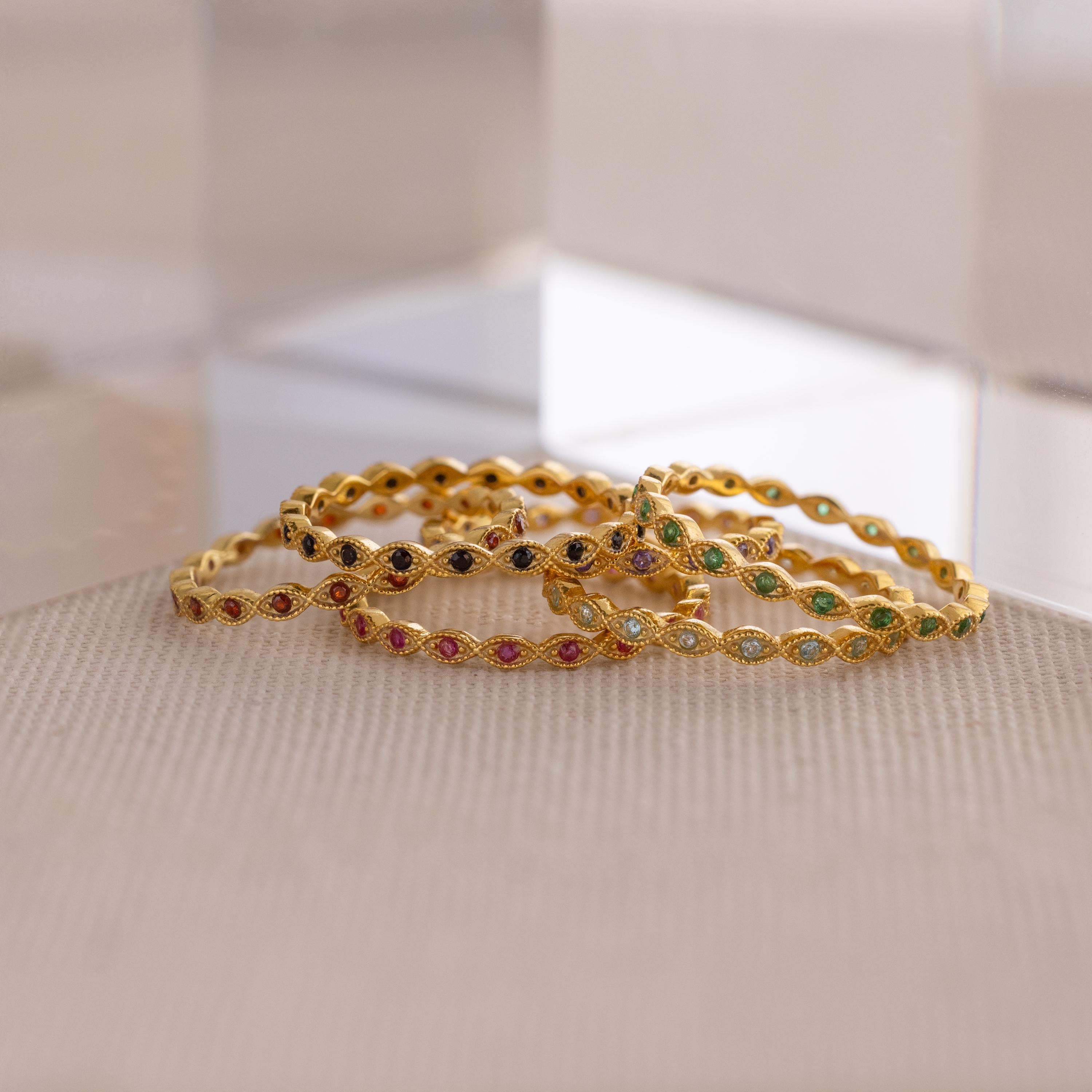 A dainty Marquise Birthstone Ring is paired with gold bangles set with colorful gemstones, all arranged on a textured beige surface near clear acrylic blocks for a touch of personalized elegance.