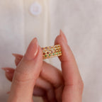A hand with manicured nails holds three stacked gold rings—including the Dainty Marquise Topaz Ring—embellished with multicolored gemstones, set against a softly blurred beige background.
