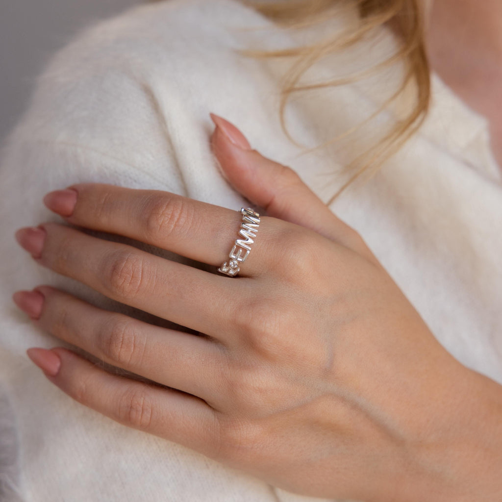 A manicured hand wears the Multiple Name Eternity Ring in Sterling Silver, personalized with "FEMINIST" in capital letters and subtle diamond accents.