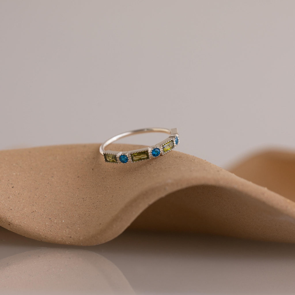 The Mixed Art Deco Birthstone Ring in Sterling Silver features green and blue stones, displayed on a curved beige surface—an ideal choice for anyone seeking a unique personalized birthstone ring.
